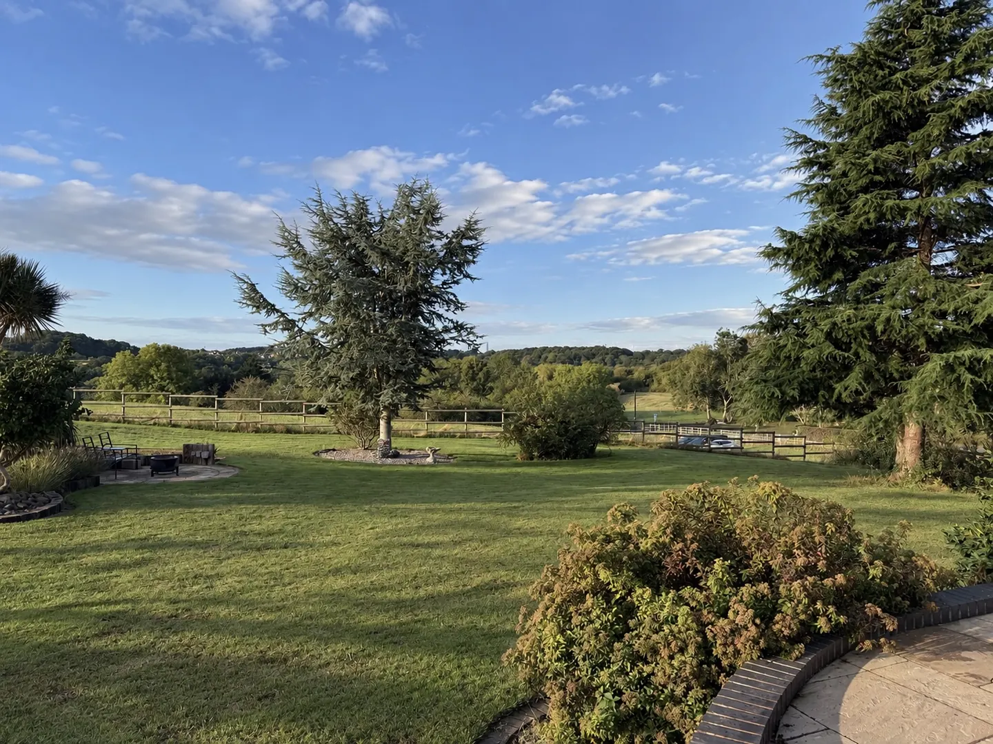 The big lawn at The Woodlands under a blue sky, tall evergreen trees and a post-and-rail fence, with wooded hills on the horizon
